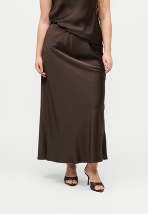 Γυναίκα που φοράει καφέ σατέν μίντι φούστα συνδυασμένη με ταίριαστα ανοιχτά ψηλοτάκουνα παπούτσια σε απλό φόντο.