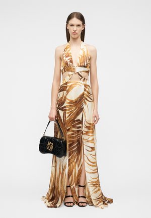 Kvinde iført brun og beige tropical-print halterneck-kjole med udskæringer, holder sort quiltet håndtaske og har sorte sandaler med remme på.