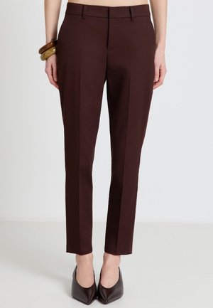 Schmal geschnittene dunkelbraune Hose, getragen mit dunklen spitzen Absätzen, accessorisiert mit auffälligen Armbändern am linken Handgelenk.