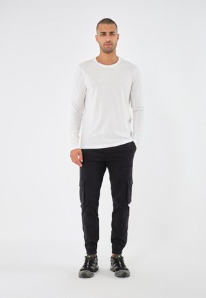 T-shirt blanc à manches longues, pantalons cargo noirs avec poches latérales, et baskets noires avec des accents verts. Fond uni mettant en valeur les détails de la tenue.