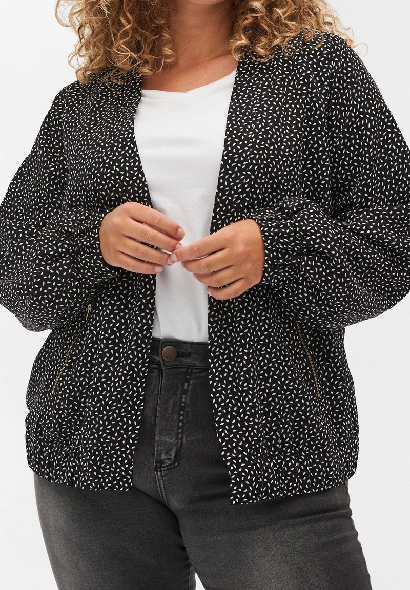Femme aux cheveux bouclés porte une veste à motif noir et blanc par-dessus une chemise blanche et un jean taille haute gris foncé, debout les mains près de la veste.