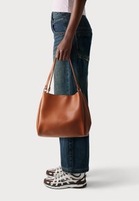 Sac à main en cuir marron avec une finition lisse, une forme arrondie et des bandoulières. Porté avec un pantalon en denim bleu et des baskets marron et blanches.