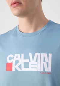 Šviesiai mėlynas medvilnės marškinėlis su apvalia iškirpte. Pabrėžia ryškų baltą užrašą "CALVIN KLEIN" ir raudoną kvadratą. Lygi tekstūra.