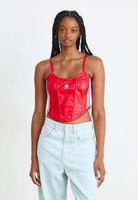 Raudonas latekso trumpas viršus su plonomis petnešėlėmis, puošiamas baltu Adidas logotipu ir baltomis šoninėmis juostelėmis, derinamas su šviesiai mėlynomis aukšto liemens džinsomis.