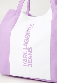 Lila och vit tygväska med texten "KARL LAGERFELD JEANS" i vertikal inriktning. Tillverkad av hållbart material, med två axelremmar.