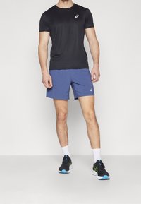 Svart kortärmad träningsskjorta, blå shorts, vita strumpor, svarta och blå löparskor med gröna detaljer, modell som står mot en neutral bakgrund.