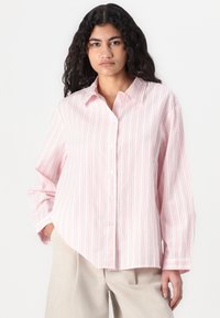 Camicia a righe con bottoni di colore rosa chiaro e linee verticali bianche, vestibilità comoda, maniche lunghe e tessuto di cotone liscio. Dettaglio della tasca frontale.