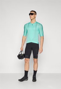Ein Mann trägt ein mintgrünes Radtrikot und schwarze Shorts, während er einen schwarzen Helm hält. Das Trikot hat einen Reißverschluss und ein Logo. Schwarze Radschuhe vervollständigen das Outfit.