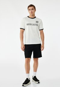 Biała koszulka z krótkim rękawem z czarnym wykończeniem z napisem "10 MIAMI SOCCER CULTURE", w połączeniu z czarnymi szortami i czarnymi sneakersami.