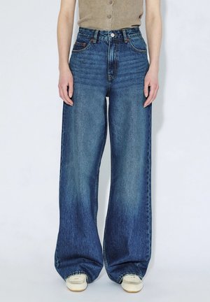 Osoba nosząca niebieskie jeansy z wysokim stanem i szerokimi nogawkami, połączone z beżową bluzką i białymi trampkami, stojąca na tle jednolitego jasnego tła.