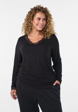 Lächelnde Frau mit lockigem Haar, die ein schwarzes Langarmtop mit Spitzenbesatz und schwarze Hose trägt, steht mit einer Hand in der Tasche.