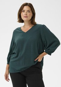 Femme portant une blouse à manches longues texturée vert foncé avec un col en V et un pantalon noir, posant avec une main dans la poche sur fond blanc.