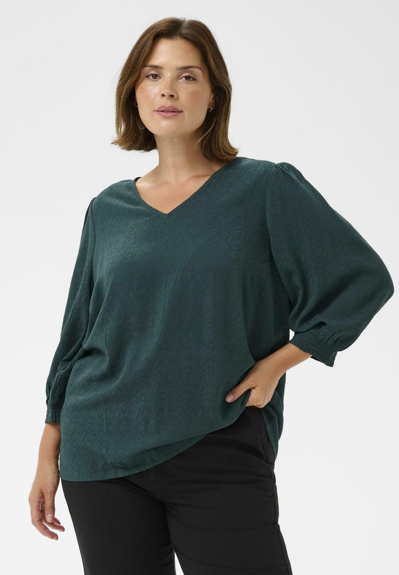 Femme portant une blouse à manches longues texturée vert foncé avec un col en V et un pantalon noir, posant avec une main dans la poche sur fond blanc.