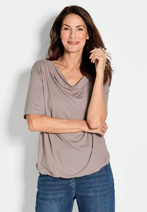 Cowl-Neck, Kurzarm-Top aus leichtem Taupe-Stoff mit drapiertem Design, kombiniert mit blauen Jeans. Weiche Textur, lockere Passform.