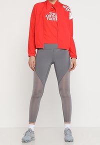 Röd lättviktjacka med logotyp, kombinerad med figurnära grå leggings med strukturerade paneler. Grå träningsskor fulländar looken.
