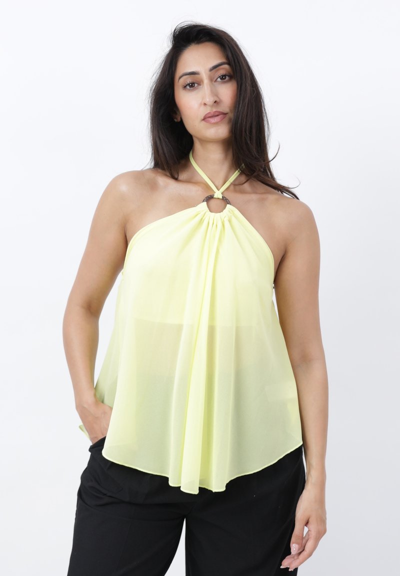 Mujer con cabello largo y oscuro lleva un top halter amarillo pálido sin mangas con detalle de anillo, combinado con pantalones negros sobre fondo blanco liso.