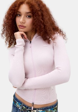 Ung kvinde med krøllet rødt hår iført en lyserød ribstrikket cardigan med lynlås og blå jeans med et mønstret linning, poserer tankefuldt.