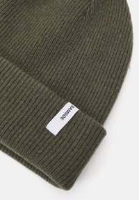Žalias megztas beanie su raukšlėtu tekstūru, sulenkta apvadu, kuriame yra balta etiketė su "SAMSOE" tekstu. Minkšta medžiaga, universalus dizainas.