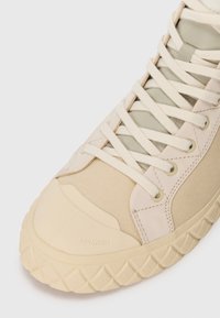 Baskets montantes beige avec semelle en caoutchouc texturé, tige en toile de coton et accents vert clair contrastants. Présente des lacets ronds et des œillets en métal.