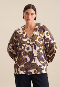Blusa stampata in color crema con motivi paisley blu e oro. Presenta una scollatura a V profonda e maniche a palloncino. Tessuto dalla texture liscia.