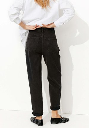 Personne portant un jean noir retroussé et des chaussures noires à enfiler cloutées, debout contre un mur blanc avec les mains derrière le dos.