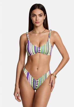 Donna che indossa un bikini a righe viola, verdi e bianche con spalline arricciate, collana d'oro e braccialetto d'oro, in piedi davanti a uno sfondo semplice.