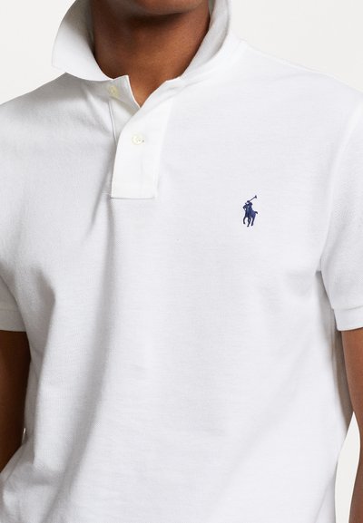 Nahaufnahme einer Person, die ein weißes Polohemd mit aufgestelltem Kragen und einem kleinen dunkelblauen Polo-Spieler-Logo auf der linken Brust trägt.