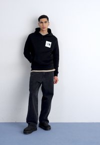 Felpa nera con logo frontale, abbinata a una maglietta beige a strati, jeans neri larghi e scarpe nere. Tessuto morbido e design casual.