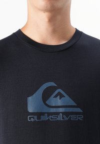 Artimas vyro liemens kadras, vilkintis juodą marškinėlį su mėlynu Quiksilver logotipu, pabrėžiantis iškirptę ir logotipo dizainą.