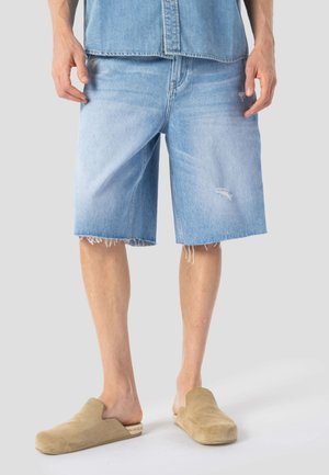 Hellblaue Jeansshorts mit weitem Schnitt, ausgefranstem Saum und kleinen Used-Details. Kombiniert mit beigen Slippern.