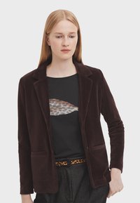 Blazer in corduroy marrone con colletto a rever, due tasche frontali e tessuto testurizzato; indossato sopra una camicia nera con una grafica di pesce.