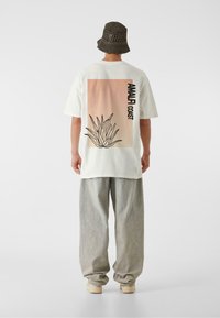 Cămășea albă oversized prezintă un grafic bej cu o contur al unei plante de agave și textul "AMALFI COAST". Este purtată cu pantaloni gri lejeri și o pălărie de tricot.
