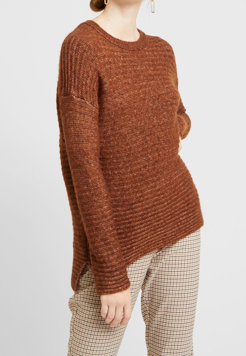 Brauner gestrickter Pullover mit geripptem Muster, Rundhalsausschnitt, seitlichen Schlitzen und lockerer Passform, getragen über karierten Hosen.