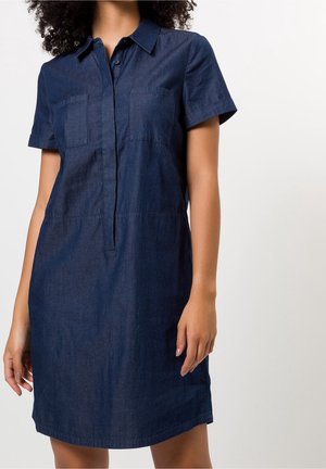 Donna che indossa un abito camicia in denim blu scuro a maniche corte, con colletto e tasche frontali, su uno sfondo semplice.