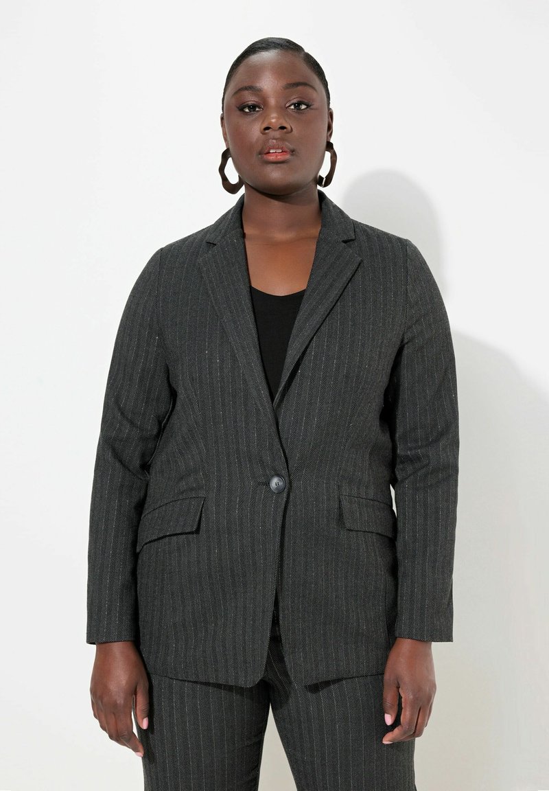 Grijze krijtstreep blazer met een enkele knoop, twee voorzakken en een getailleerde pasvorm. Draagt over een zwart topje, gecombineerd met bijpassende onderkant.