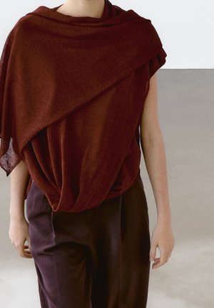 Personne portant un haut asymétrique ample de couleur rouge profond associé à un pantalon taille haute marron foncé, debout devant un fond uni.