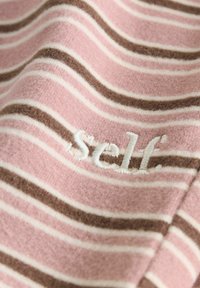 Material moale cu dungi orizontale roz, maro și albe, având text brodat alb cu cuvântul „self”.