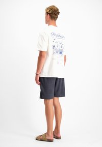 Witte t-shirt met een blauw grafisch ontwerp op de achterkant, gecombineerd met donkere gebloemde shorts en beige sandalen. Korte mouwen, ontspannen pasvorm.