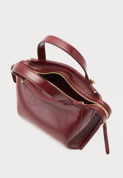 Sac à main en cuir rouge foncé avec fermeture éclair dorée partiellement ouverte et deux poignées sur fond blanc.