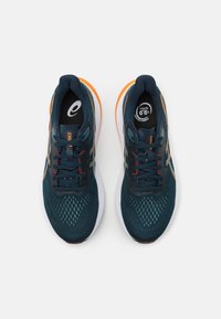 Zapatillas de running con parte superior de malla en un verde azulado oscuro, acentos naranjas, cordones negros y suela blanca. Presentan un logo en la lengüeta y una textura con patrón.