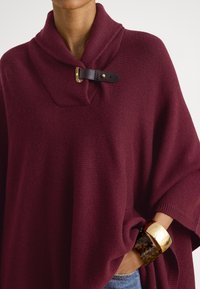 Γυναίκα που φοράει μπορντό πλεκτό πόντσο με δερμάτινη αγκράφα στο γιακά και χρυσά και χελώνα βραχιόλια στον καρπό της.