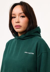 Grüne Baumwoll-Hoodie mit lässiger Passform, auf der Vorderseite mit dem Text "DAILY COTTON" in Weiß. Weiche Textur und minimalistisches Design.