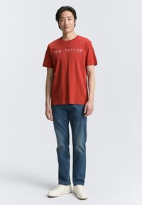 Camiseta de algodón roja con el logo "TOM TAILOR", combinada con jeans de mezclilla azules y zapatillas blancas; mangas cortas y escote redondo.