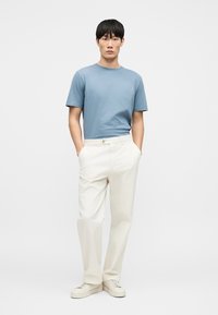 T-shirt en coton bleu clair, pantalon crème, coupe décontractée. Le mannequin porte des baskets blanches, les mains dans les poches, sur un fond neutre.