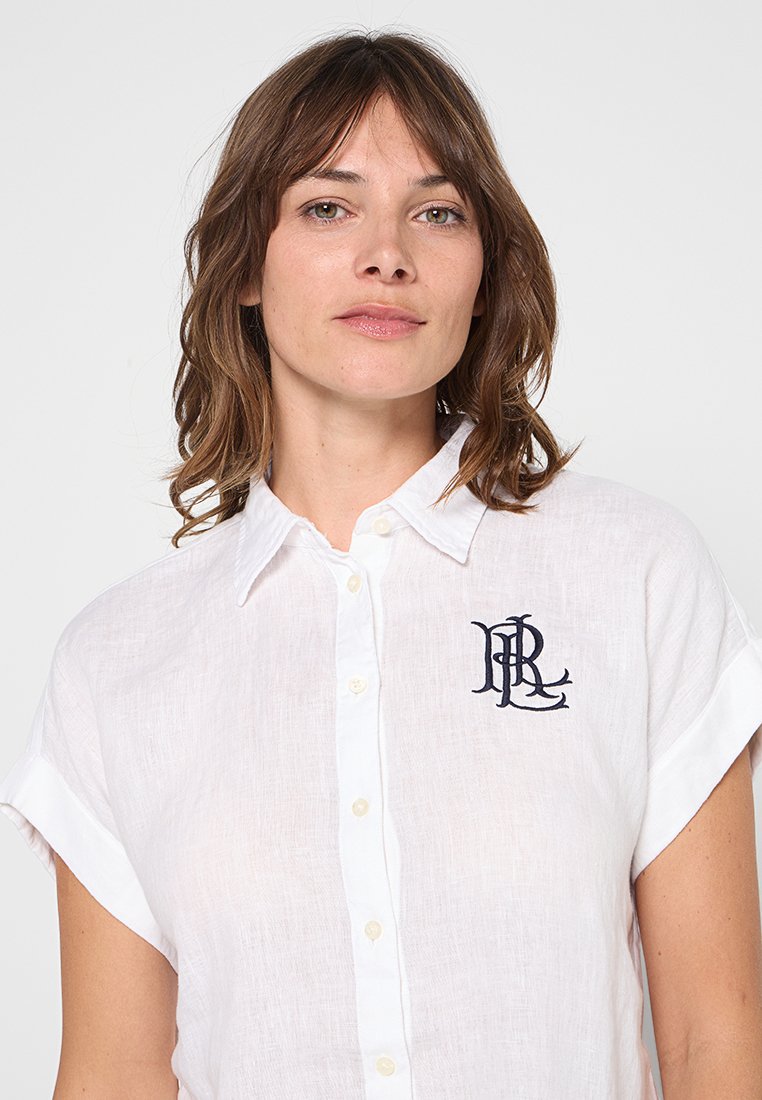 Camicia bianca di lino con maniche corte, caratterizzata da un logo ricamato blu navy sul lato sinistro del petto e una patta con bottoni sul davanti.