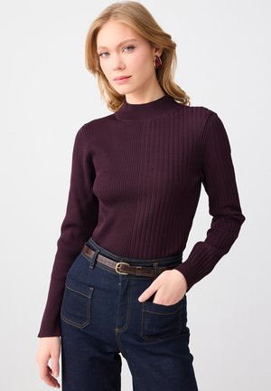 Blonde vrouw met een donkerpaars geribd shirt met lange mouwen, donkerblauwe jeans met hoge taille en een bruine riem, en rode creolen.