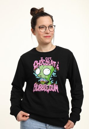 Frau mit Brille und zu einem Dutt gebundenen Haaren, die einen schwarzen Pullover mit einer grünen Comicfigur und rosa Schrift „Ich habe Schokoladen-Kaugummi.“ trägt.