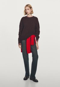 Maglione a trecce marrone scuro con scollatura tonda, abbinato a jeans scuri. Un maglione rosso legato in vita aggiunge un accento contrastante.