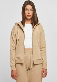Beige zip-up hettegenser med hette, frontlommer og ribbede mansjetter. Båret over en hvit topp, kombineres med beige joggebukser. Myk stoffkvalitet.