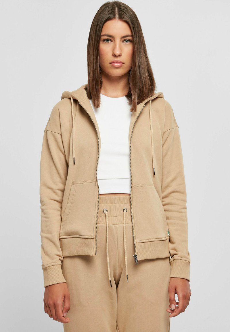 Beige zip-up hettegenser med hette, frontlommer og ribbede mansjetter. Båret over en hvit topp, kombineres med beige joggebukser. Myk stoffkvalitet.